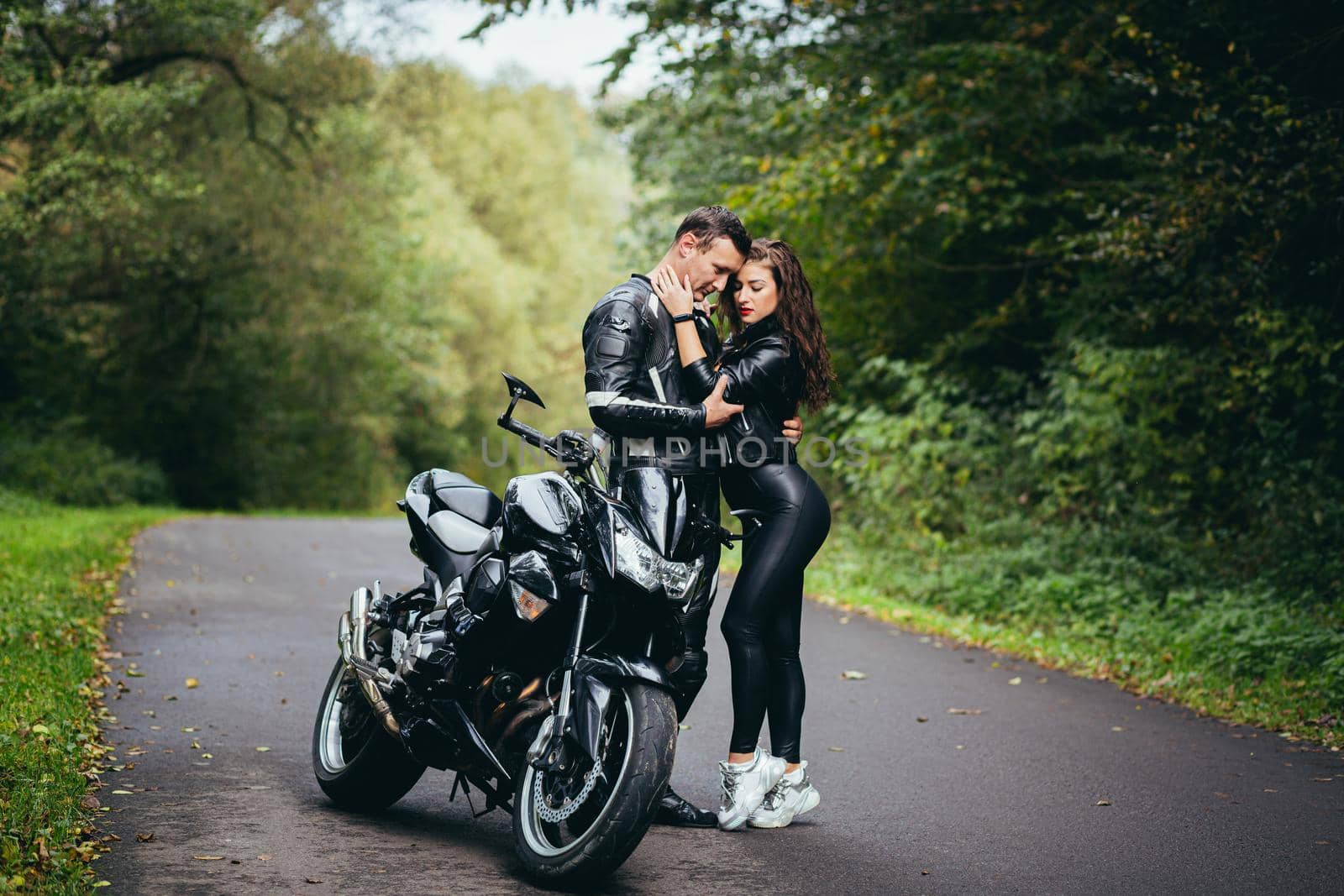 Handsome young man and beautiful young girl, couple sitting on a motorcycle, standing near a motorcycle in black leather clothes, hugging, in nature, on the street by voronaman