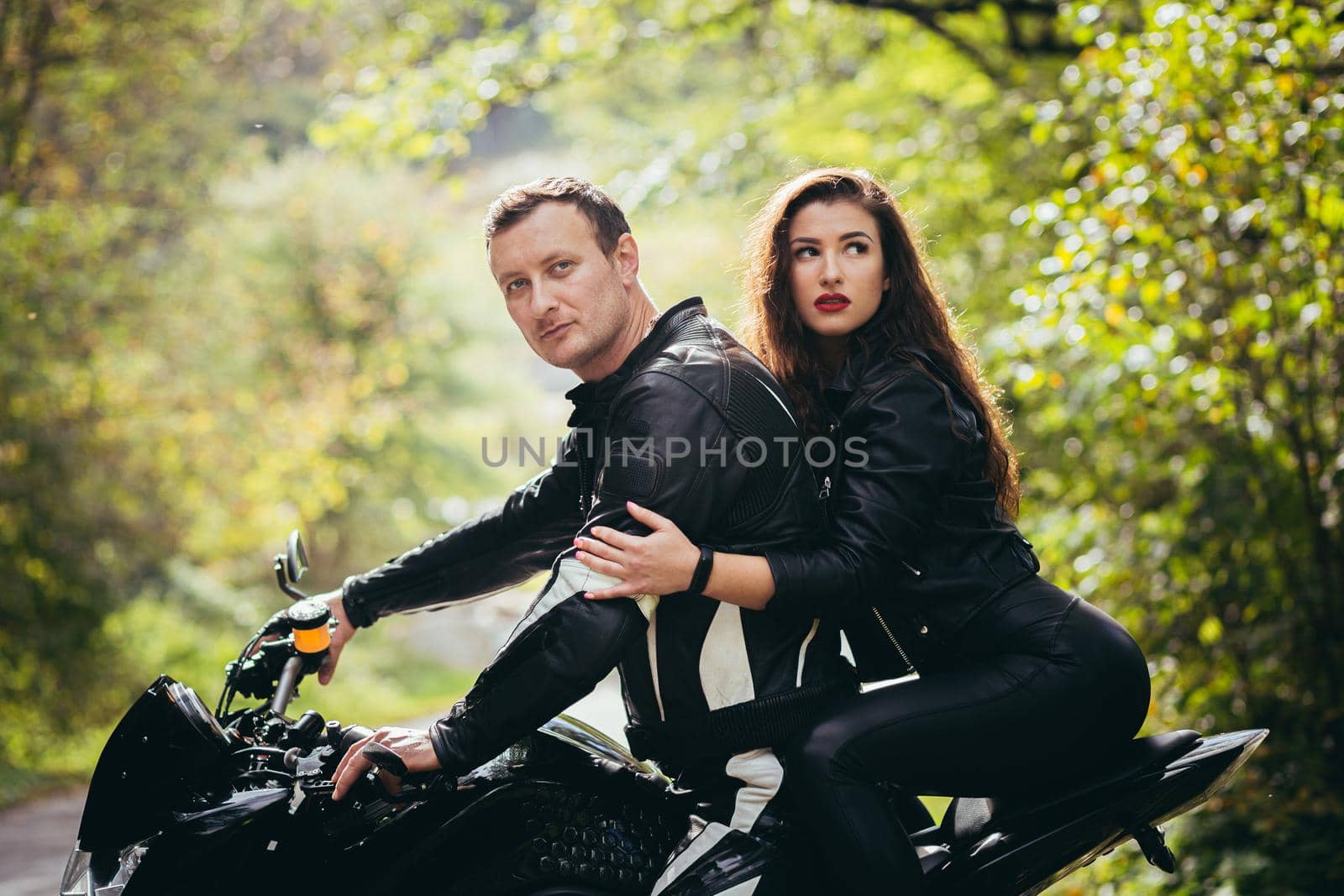 Handsome young man and beautiful young girl, couple sitting on a motorcycle in black leather clothes, close up, hugging, in nature, outdoors, looking at camera