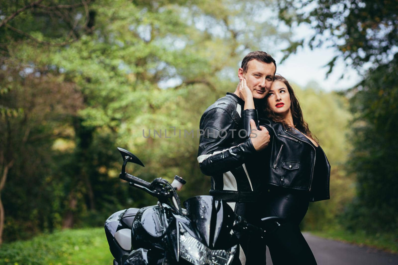 Handsome young man and beautiful young girl, couple sitting on a motorcycle, standing near a motorcycle in black leather clothes, hugging, in nature, on the street by voronaman