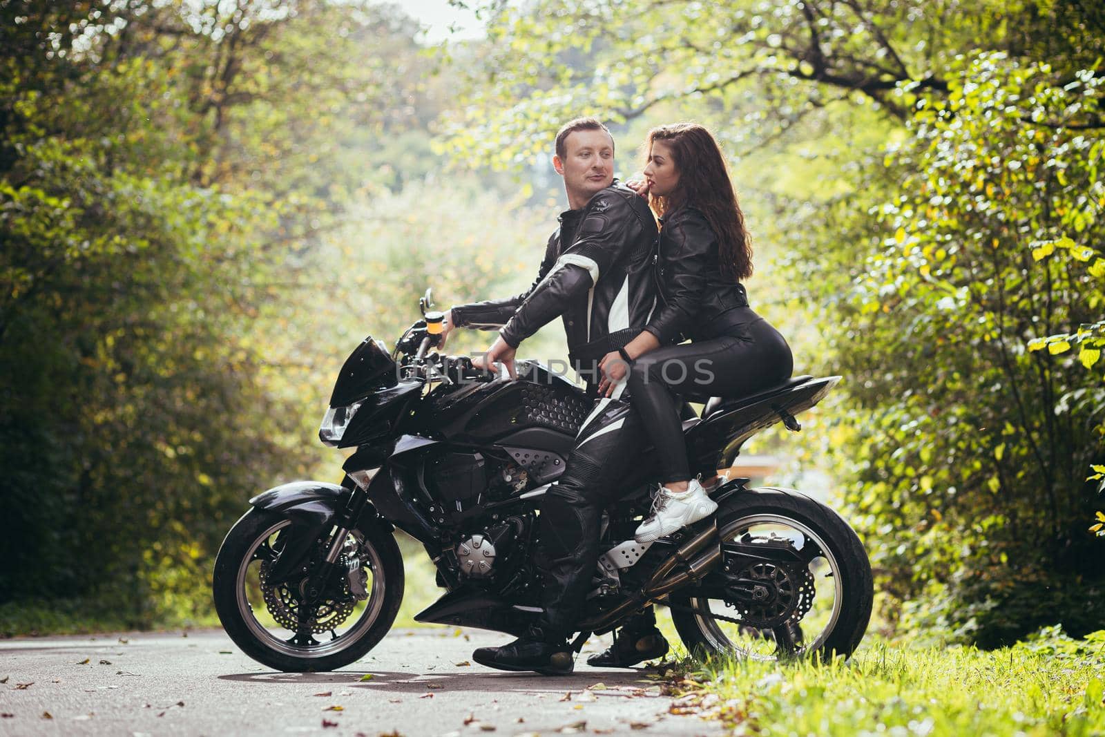 Handsome young man and beautiful young girl, couple sitting on a motorcycle, standing near a motorcycle in black leather clothes, hugging, in nature, on the street by voronaman