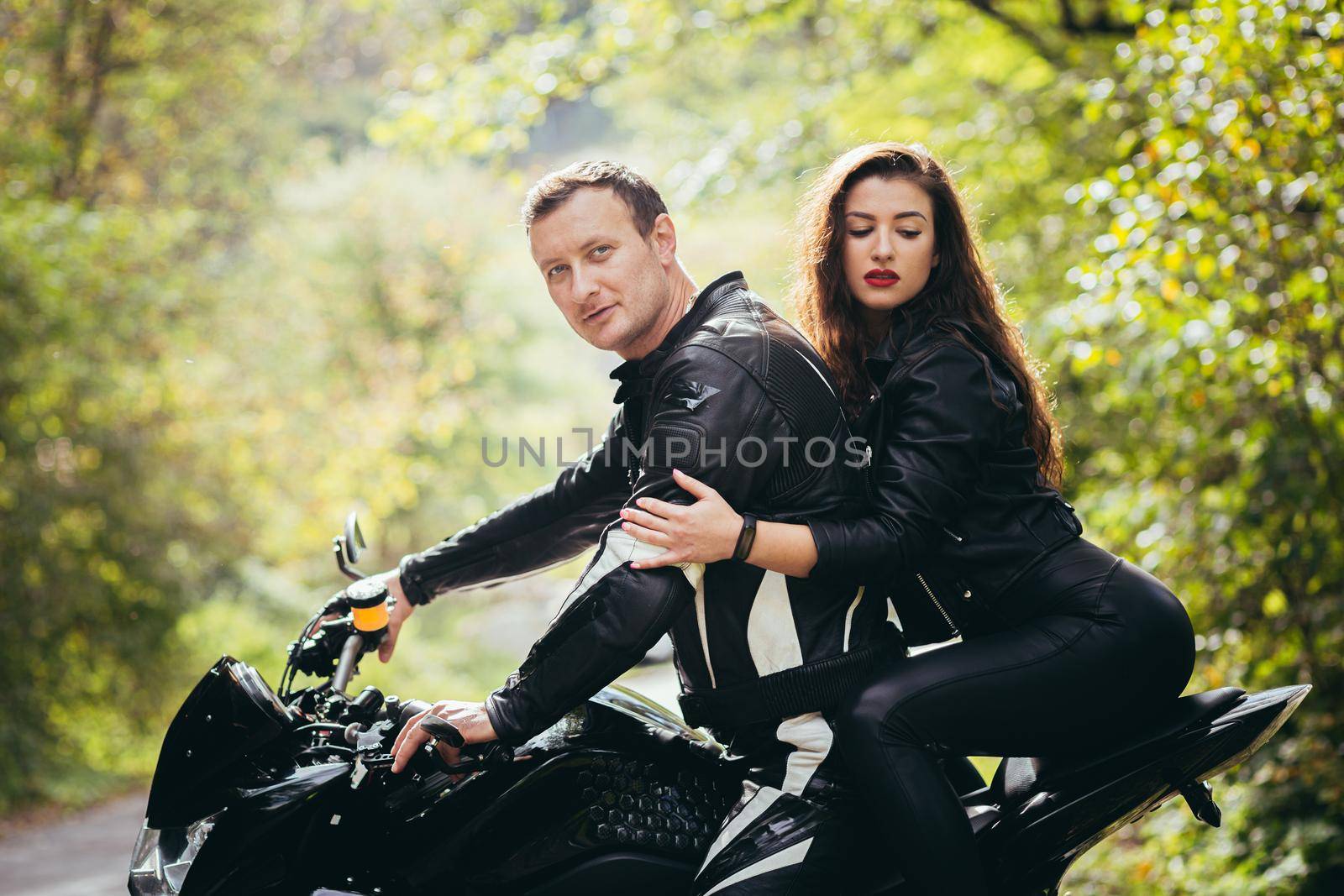 Handsome young man and beautiful young girl, couple sitting on a motorcycle in black leather clothes, close up, hugging, in nature, outdoors, looking at camera by voronaman