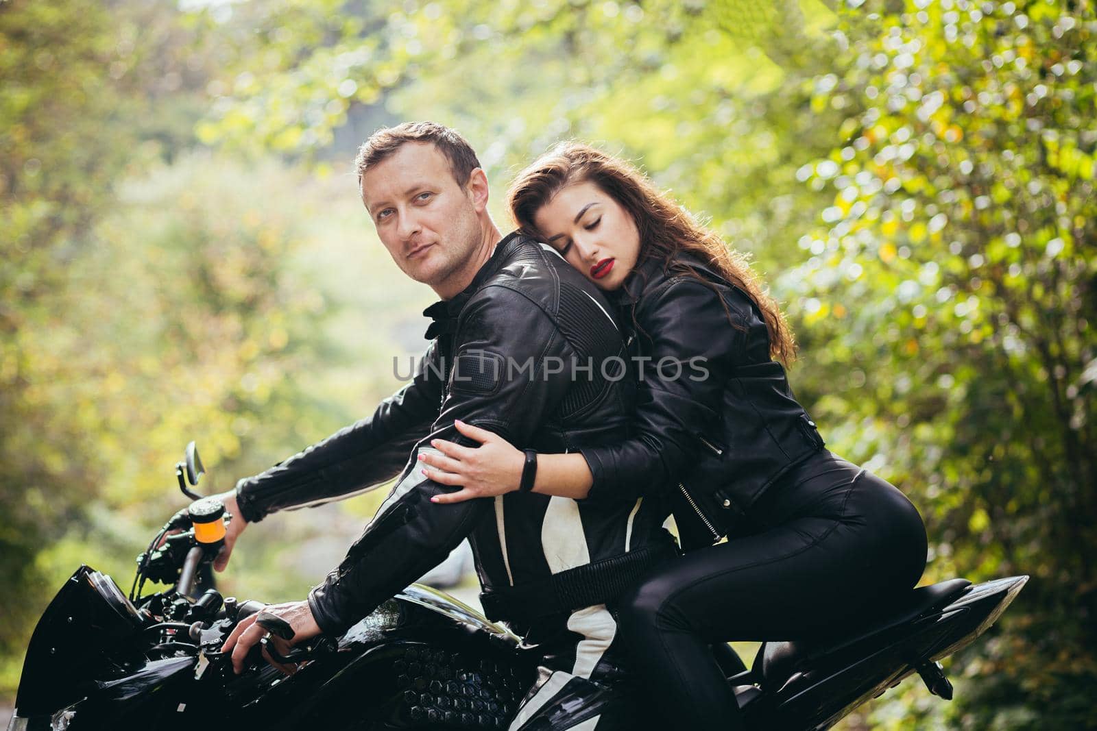 Handsome young man and beautiful young girl, couple sitting on a motorcycle in black leather clothes, close up, hugging, in nature, outdoors, looking at camera by voronaman