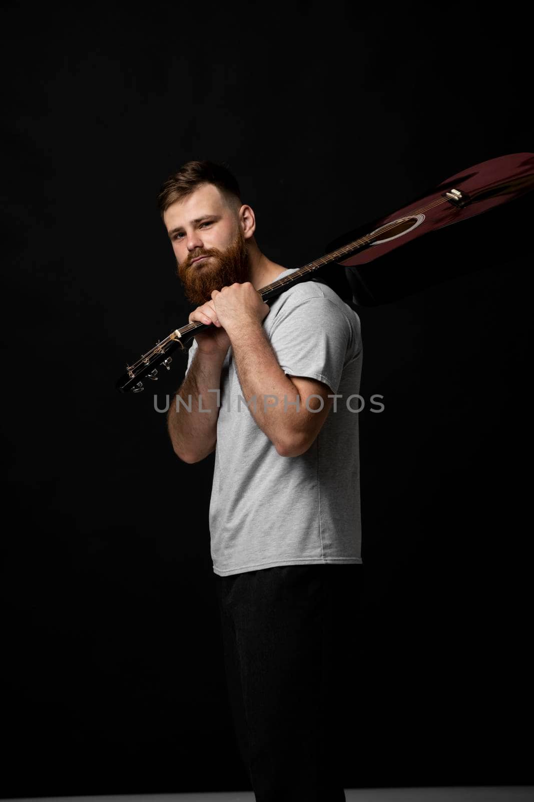 Young bearded guitarist in grey t-shirt holds a guitar on a shoulder and look in the camera in a dark room. by vovsht