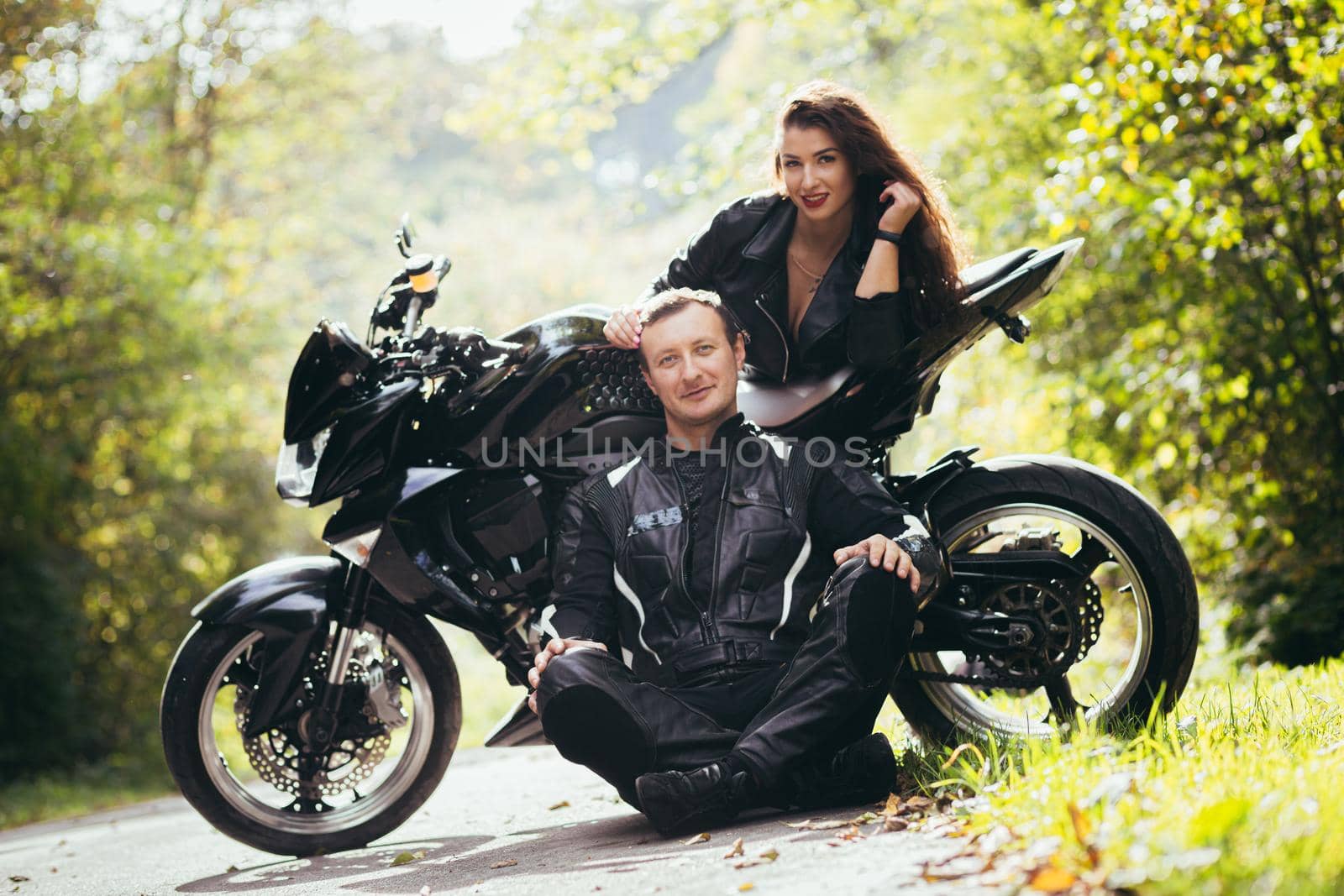 Handsome young man and beautiful young girl, couple sitting near motorcycle in black leather clothes, hugging, in nature, outdoors
