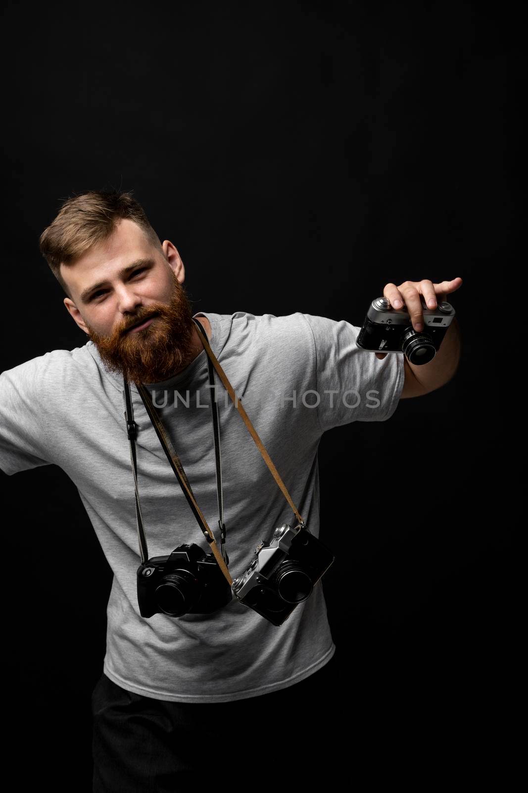 Handsome bearded photographer in a grey t-shirt with a bunch of different cameras in a hands and on a shoulder looking on a camera and ready for make a good shoot. by vovsht