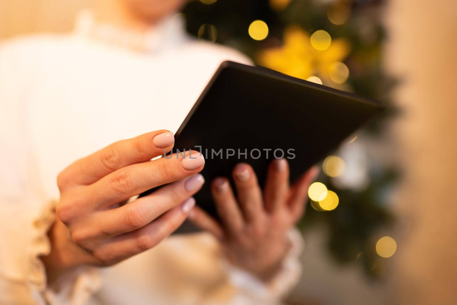 Woman in white reading interesting e-book reader while sitting near Christmas tree at home. Cozy winter pic of lady relaxing alone with novel. High quality photo