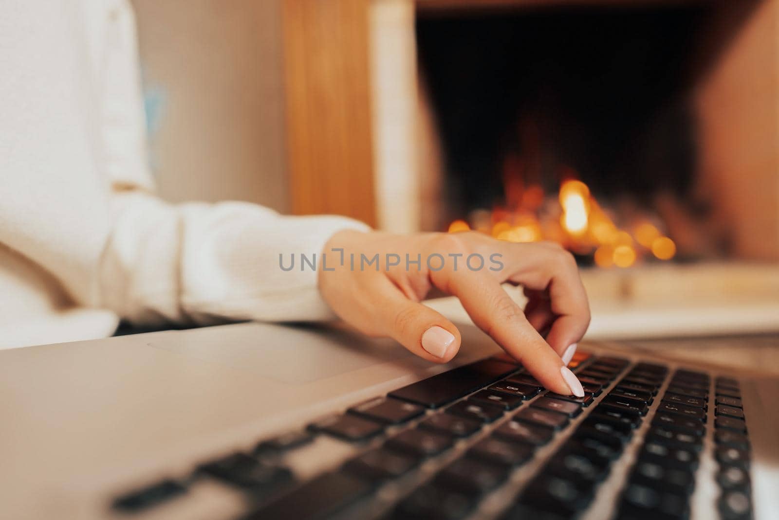 Young woman studying on laptop, sitting near cozy fireplace at home. Focus on hands typing on keyboard. Social distancing concept. Lady writing message to friends. High quality photo