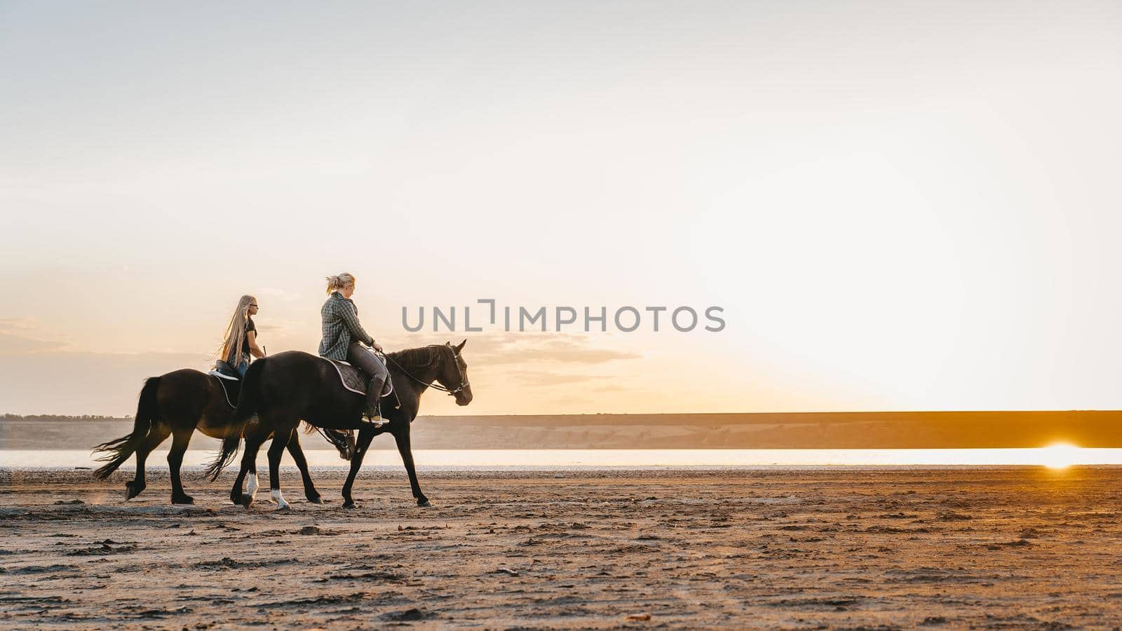 View of women riding horses along the river in golden light sunset or sunrise. Stallion walking in desert by the water