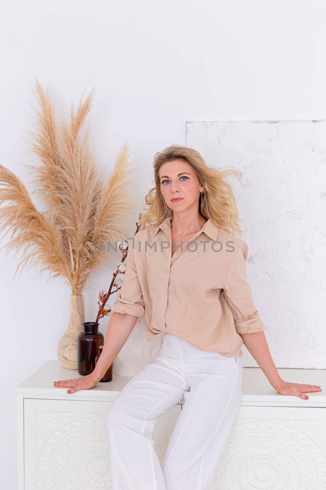 beautiful slender woman 30-40 years old with blond hair in a beige shirt and white trousers in the interior of a white room, sits on a white chest of drawers against the wall. Copy space. Vertical