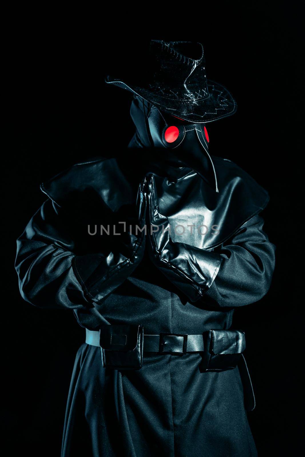 Man in plague doctor costume with crow-like mask praying with hands isolated on black background. Creepy mask, historical costume concept. Epidemic.