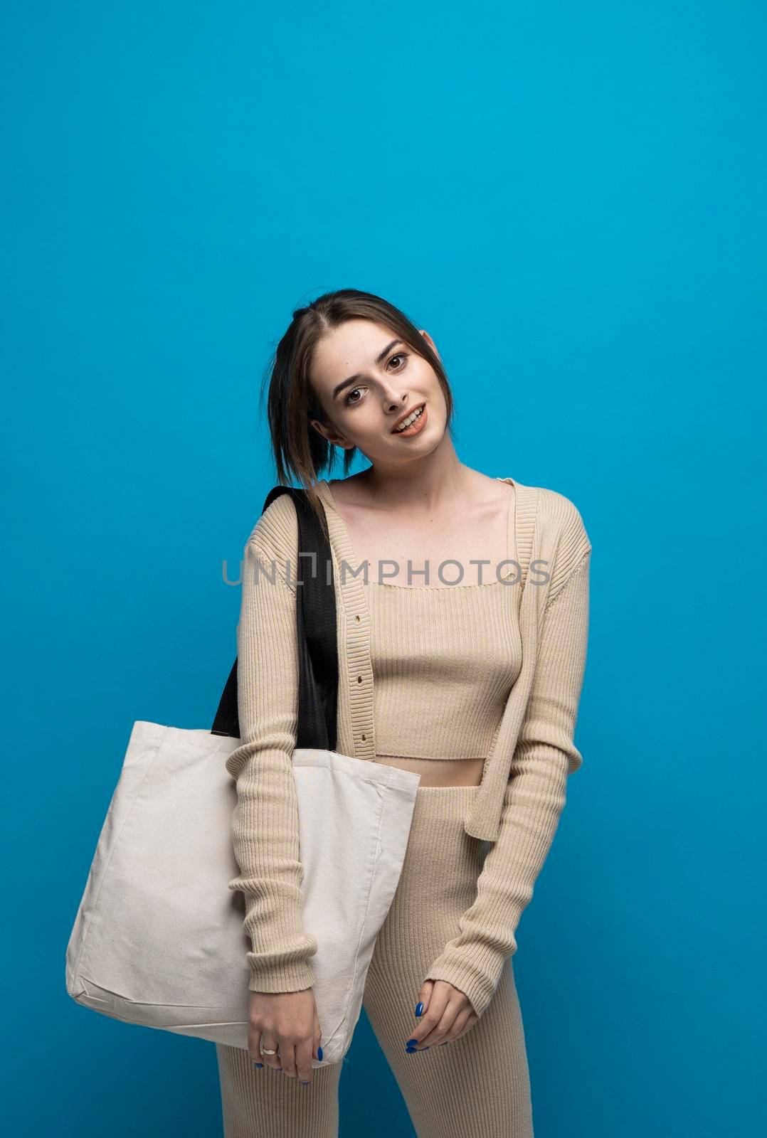 Beautiful brunette woman in a beige costume with cotton bag on a shoulder on a blue background. Mockup and zero waste concept. by vovsht