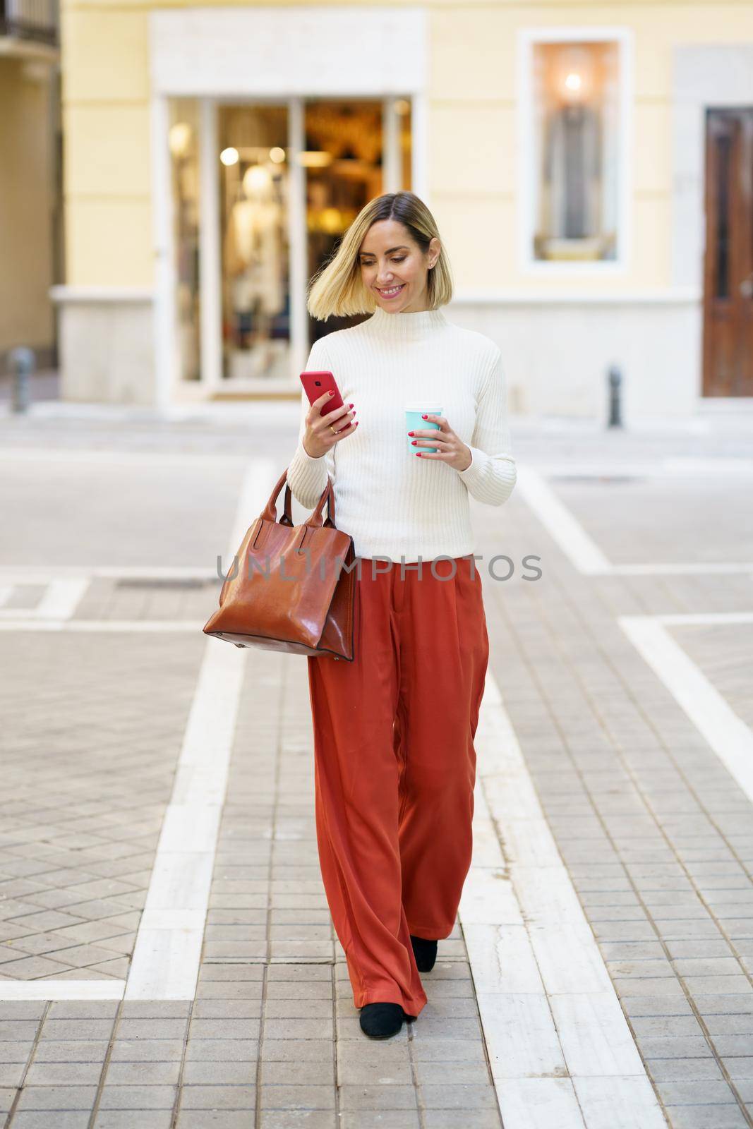 Full body of stylish female carrying handbag and cup of takeaway coffee while walking on street and using cellphone