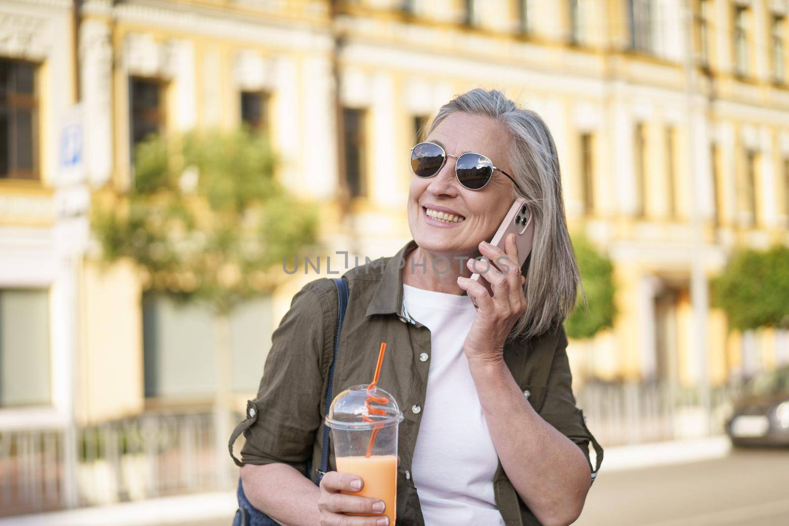 Answering phone call happy mature woman talking on the phone enjoying free time after work or traveling having juice in plastic cup on the go in city background. Enjoying life mature woman.