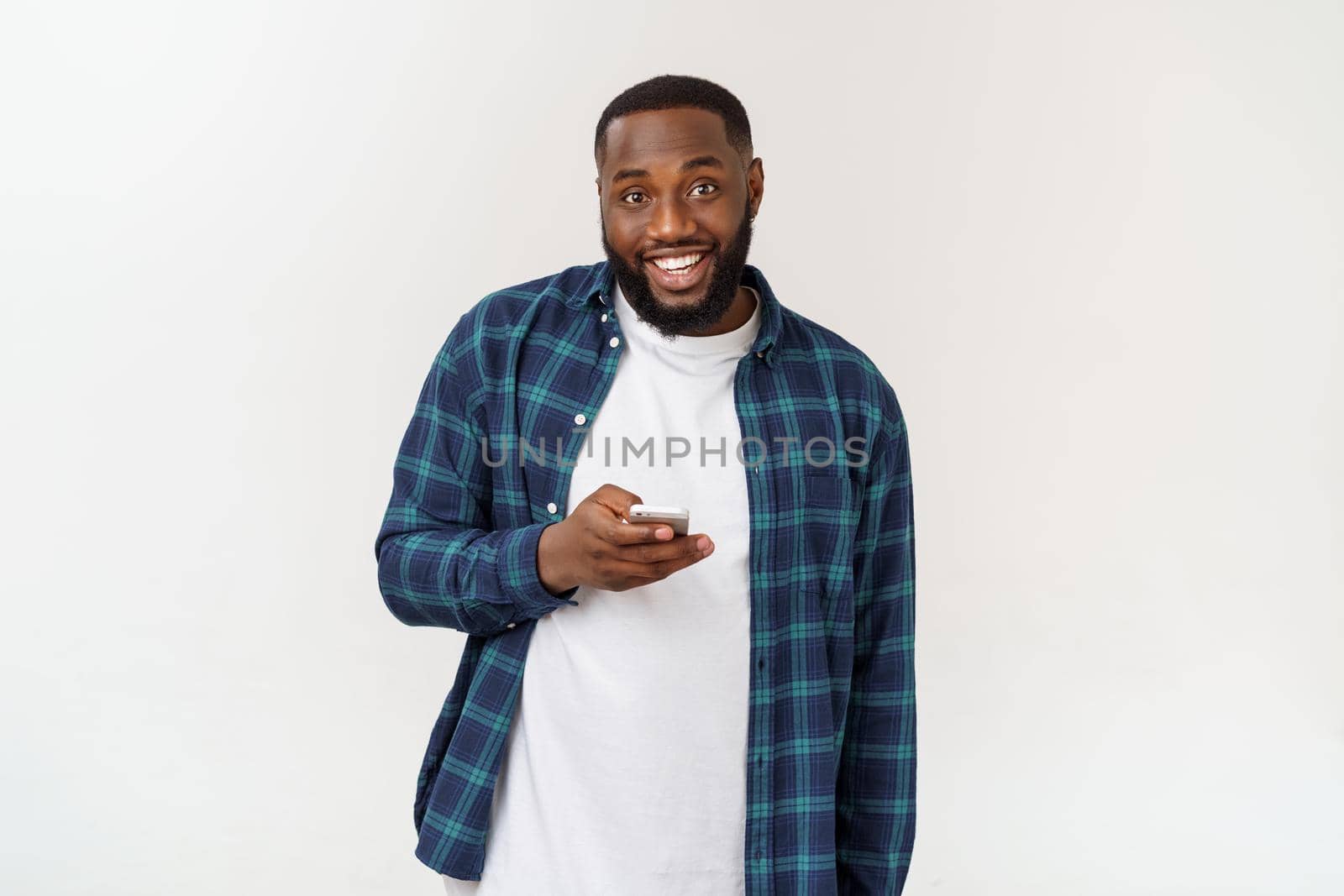 Smiling African American young man holding phone looking at smartphone isolated on white grey studio background. by Benzoix