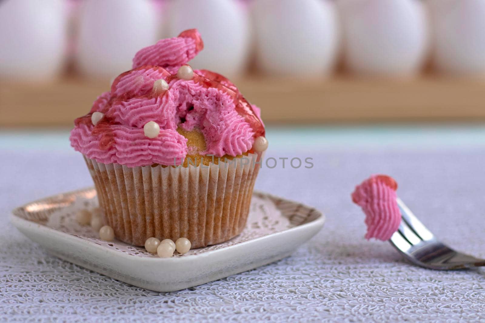 a cupcake beautifully decorated with pink cream and sugar decor stands on a square saucer on a light wooden background. A piece of muffin on a fork nearby. Sugar beads and fruit topping