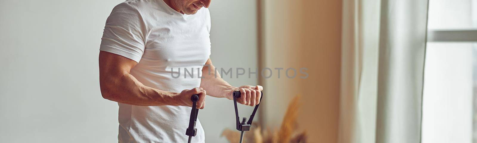 Cropped head of sporty male exercising arms with resistance bands near large window at home