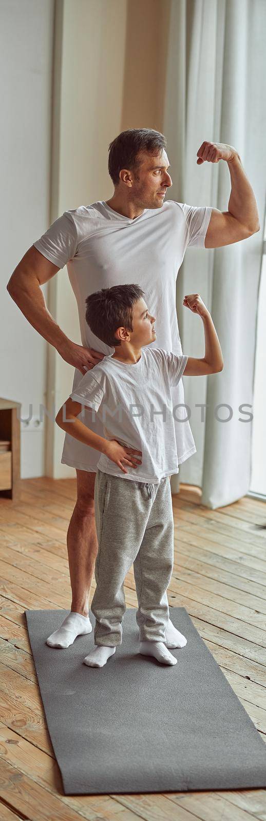 Happy muscular man and boy are standing on mat and showing biceps during domestic workout