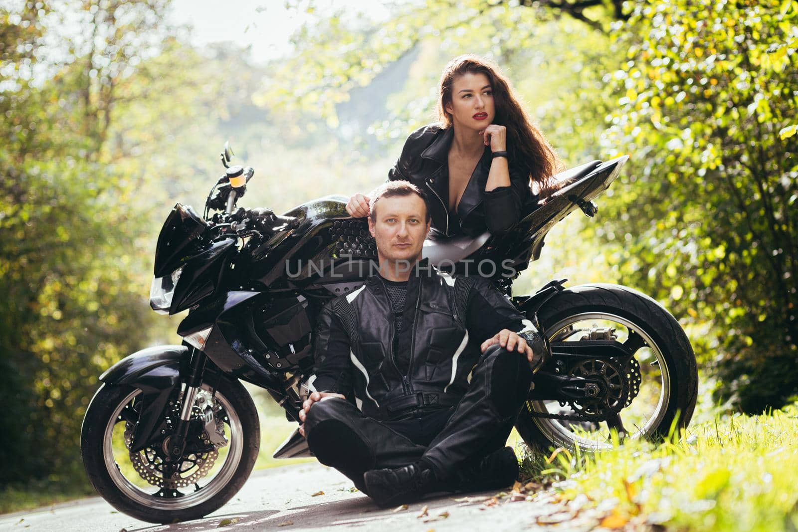 Handsome young man and beautiful young girl, couple sitting near motorcycle in black leather clothes, hugging, in nature, outdoors