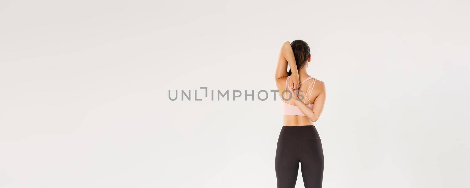 Full length rear view of active and slim brunette asian fitness girl, female athlete warm-up before yoga classes, lock hands behind back, sportswoman doing stretching exercises, white background.