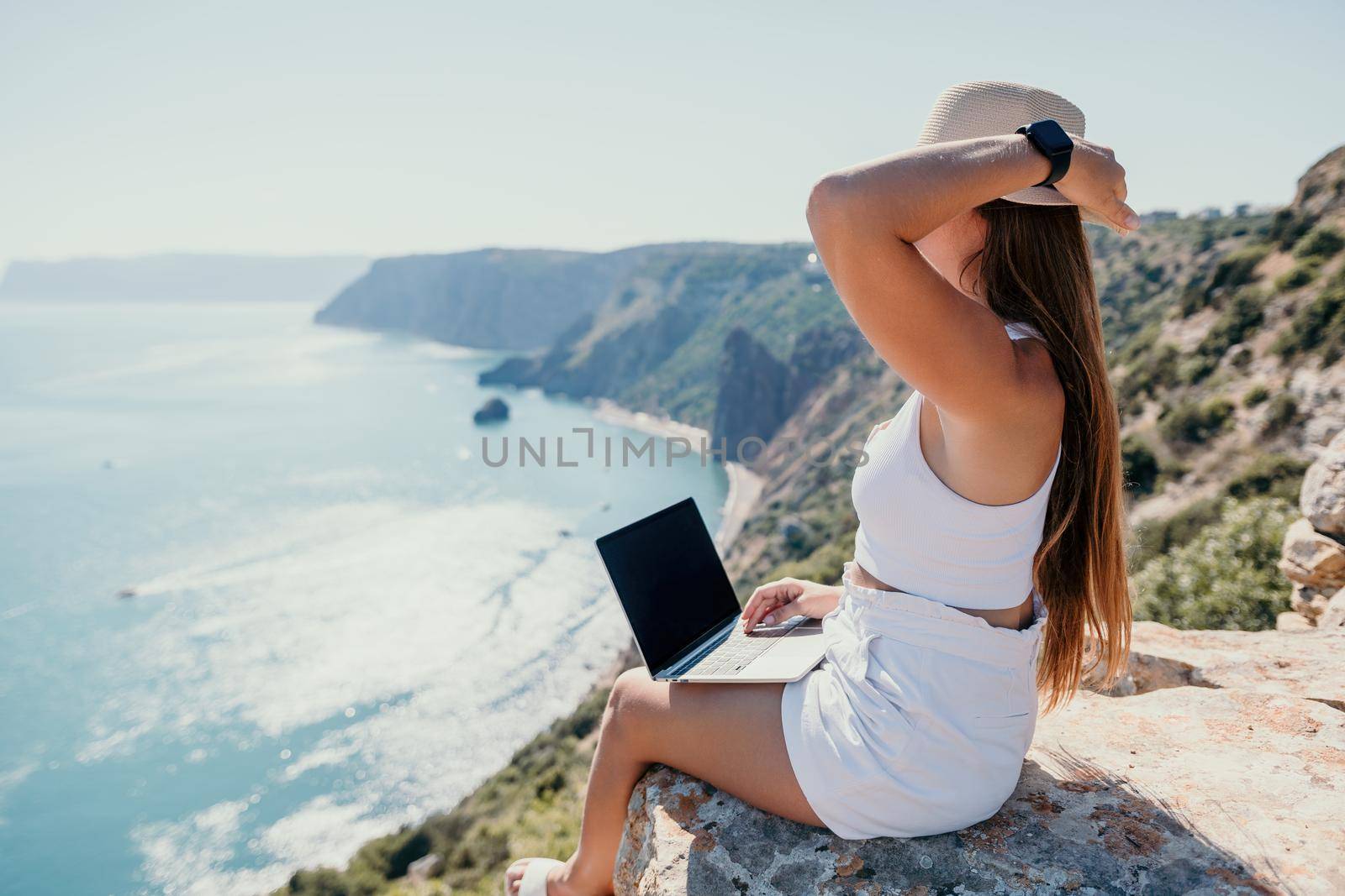 Successful business woman in yellow hat working on laptop by the sea. Pretty lady typing on computer at summer day outdoors. Freelance, travel and holidays concept.