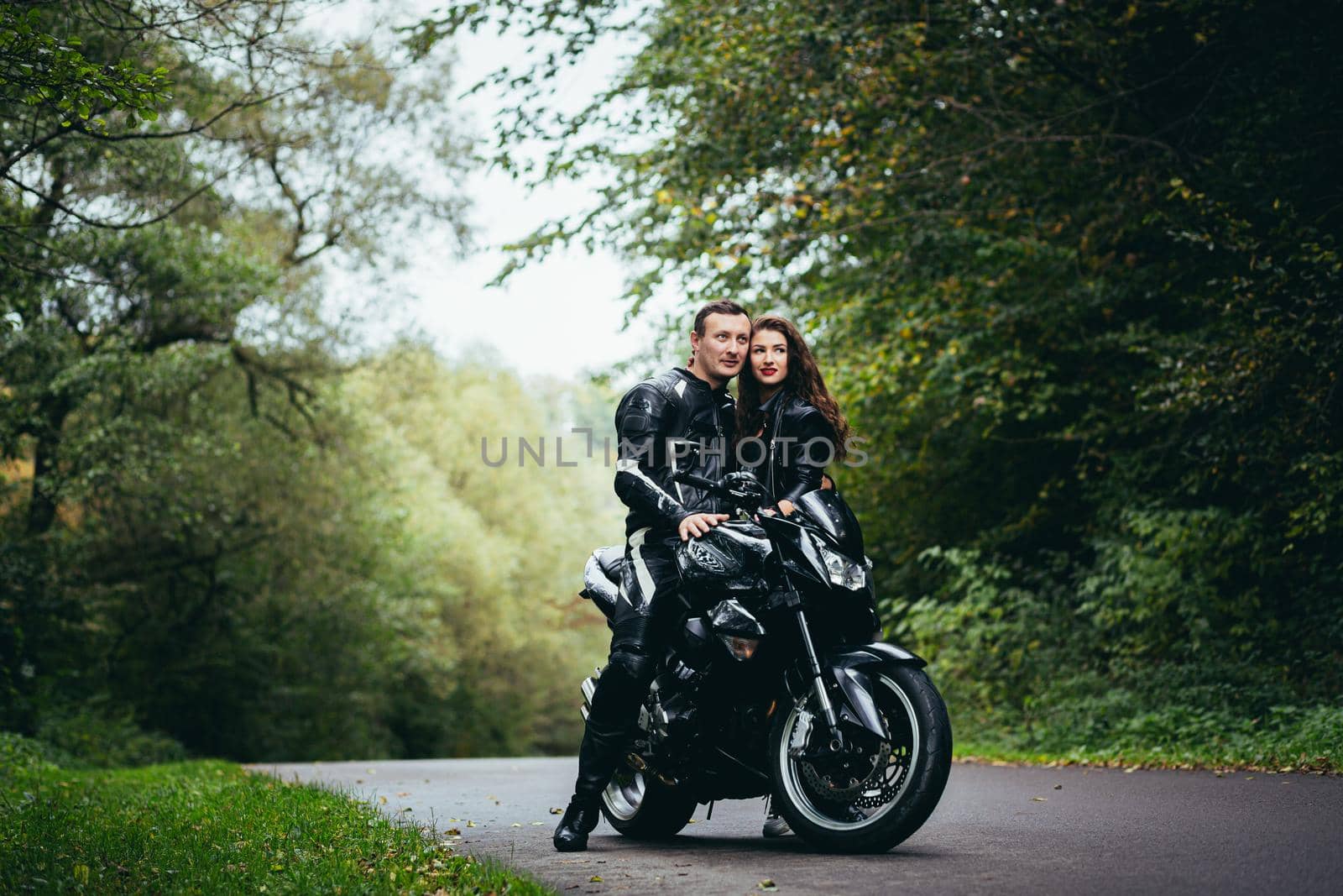 Handsome young man and beautiful young girl, couple sitting on a motorcycle, standing near a motorcycle in black leather clothes, hugging, in nature, on the street by voronaman