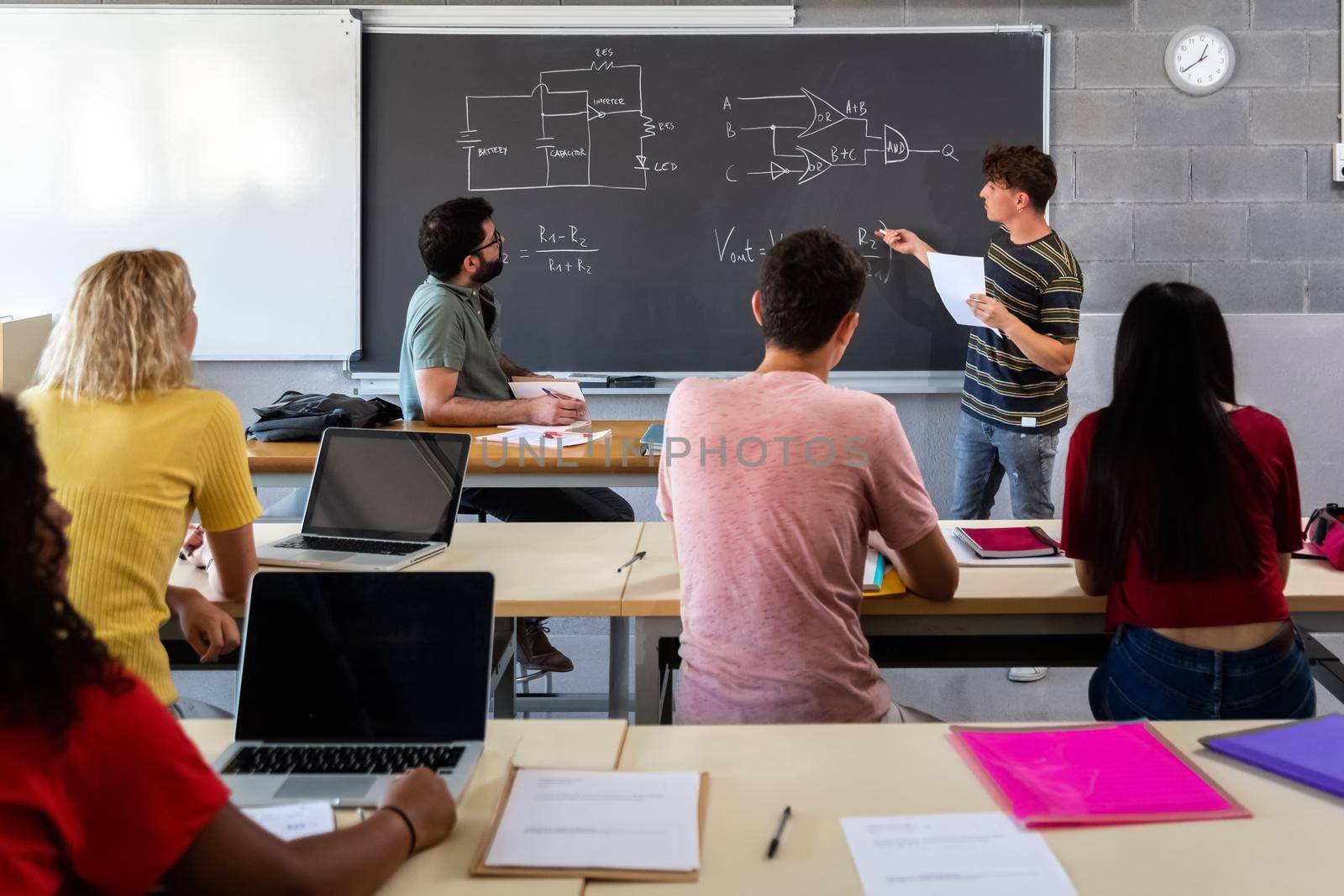 Teen male high school student giving oral class presentation in front of multiracial classmates. Teacher taking notes. by Hoverstock