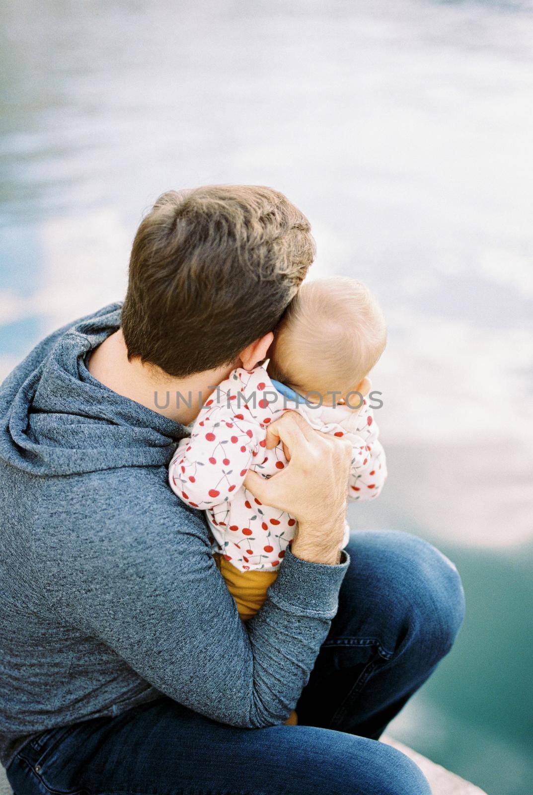 Dad sits on the pier with a baby on his lap and looks at the water. Back view. High quality photo