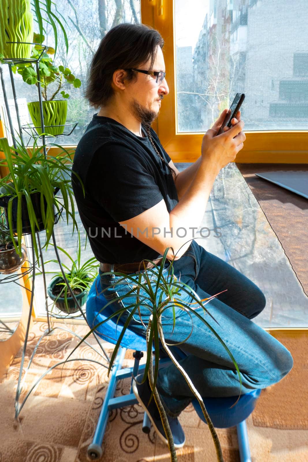 a man works at a phone in a home office, sitting on a Orthopedic ergonomic kneeling chair on a glazed balcony among greenery. back health care