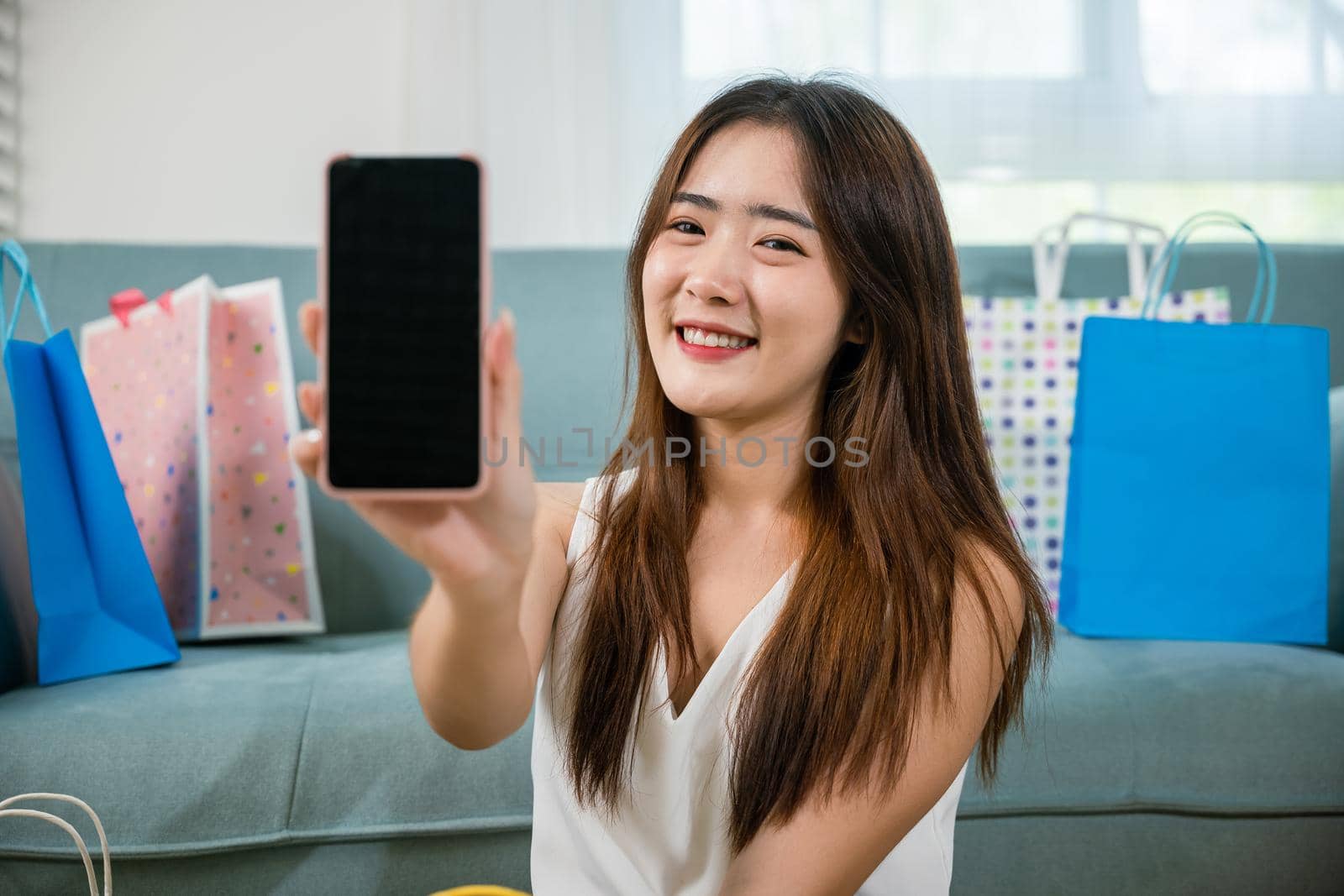 Excited Asian buying woman smiling showing mobile phone with many shopping bags at home by Sorapop