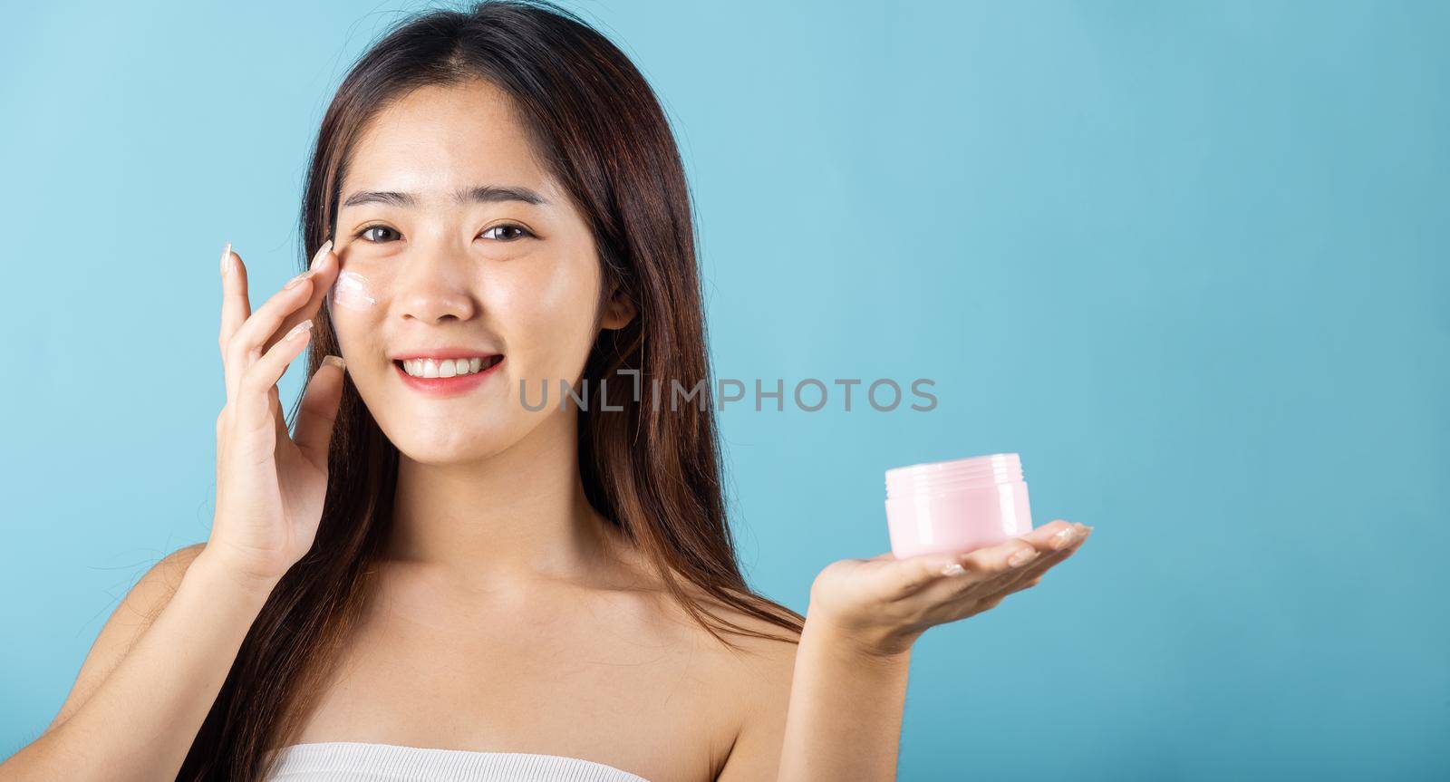 Asian young woman are not very beautiful hold jar of skin cream for face and apply anti-aging moisturizer studio isolated on blue background, Happy female pointing finger her face, pursuit of beauty