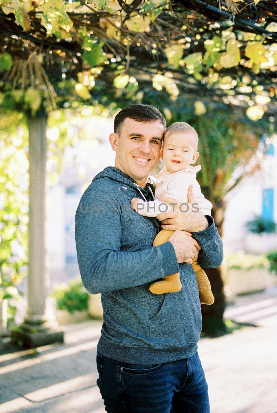 Smiling dad with a baby in his arms stands in the yard by Nadtochiy