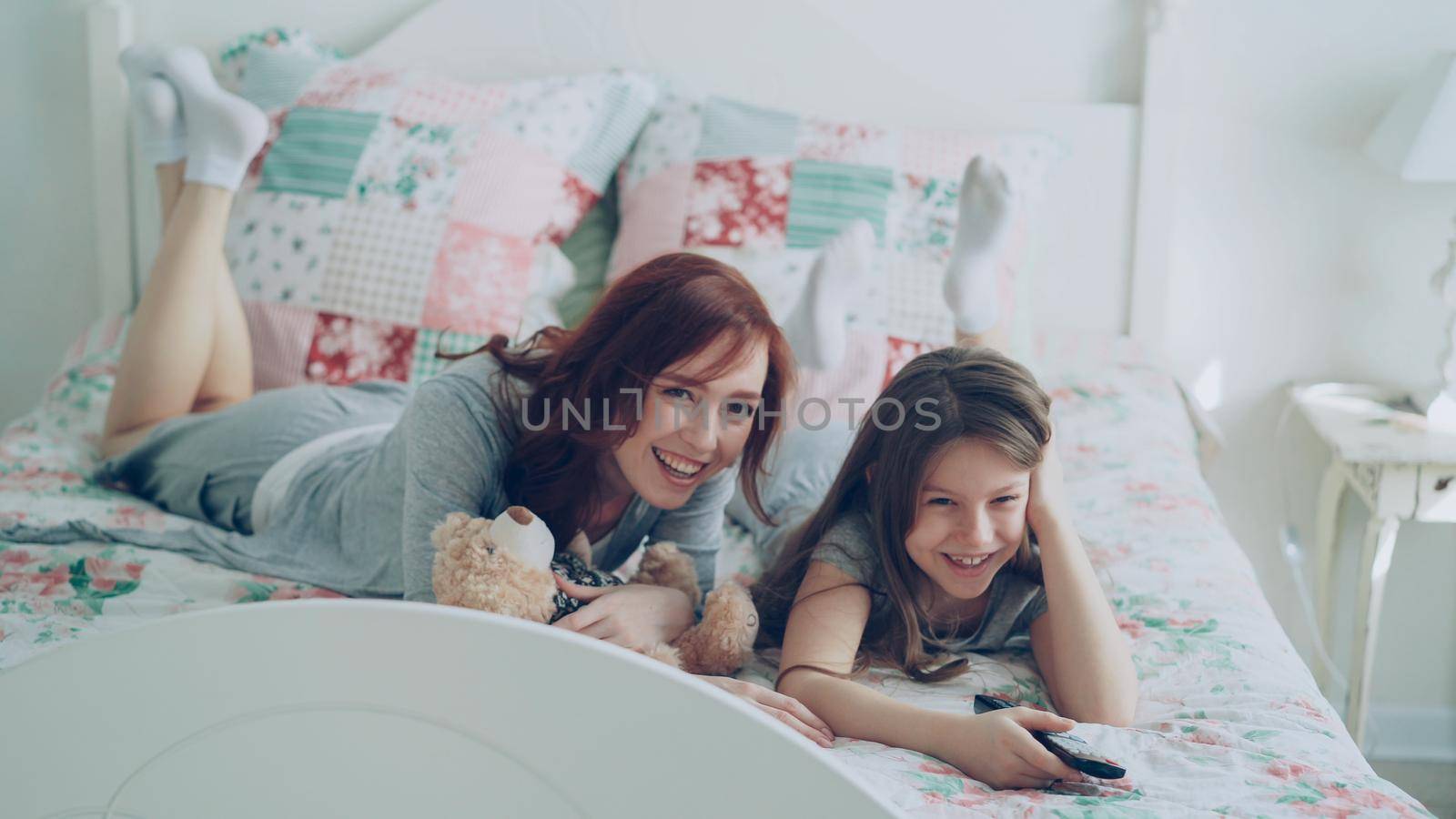 Beautiful happy mother with little daughter watching cartoon movie on TV and laughing while lying on bed at home in the morning in cozy bedroom by silverkblack