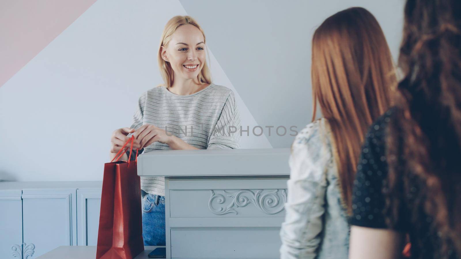 Friendly female cashier is taking garment from customer standing in line folding and packing clothes in paper bag to buyer by silverkblack