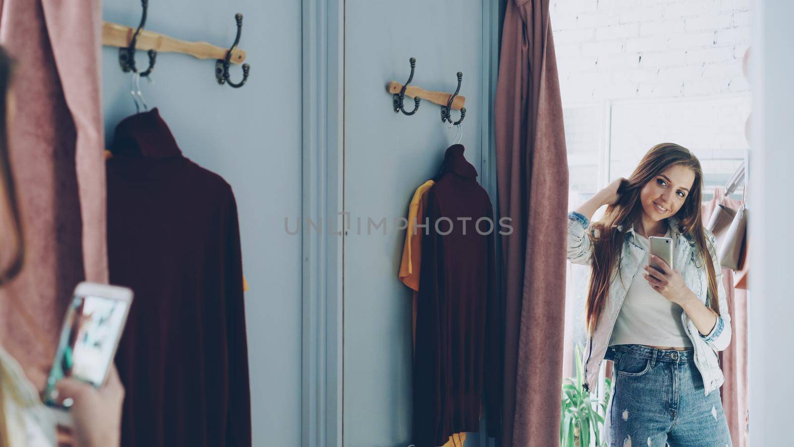 Pretty girl is making mirror selfie with smartphone while standing in fitting room in clothes store. She is posing, turning and touching her hair with lovely smile. by silverkblack