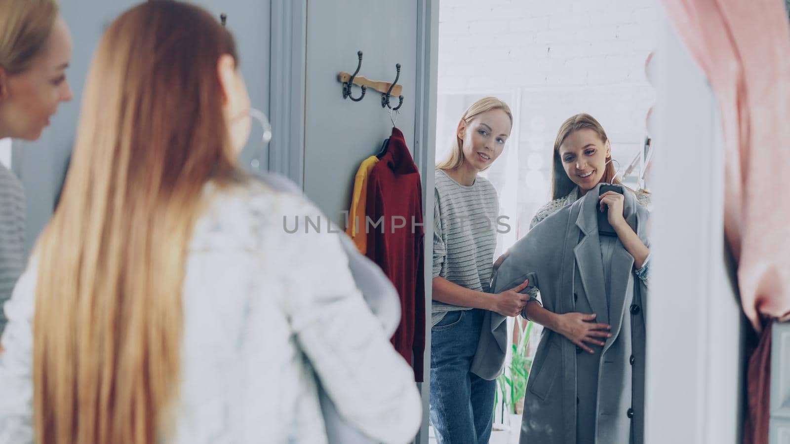 Pretty young woman is checking fashionable coat in fitting room with her friend helping her to appraise garment. They are talking, gesturing and looking at clothing in large mirror.