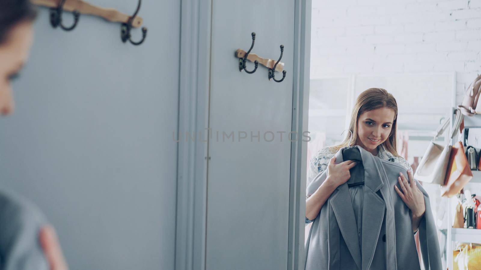 Attractive female customer is choosing stylish coat in fitting room, looking at large mirror and checking fabric and quality. She is touching garment with delight and smiling.