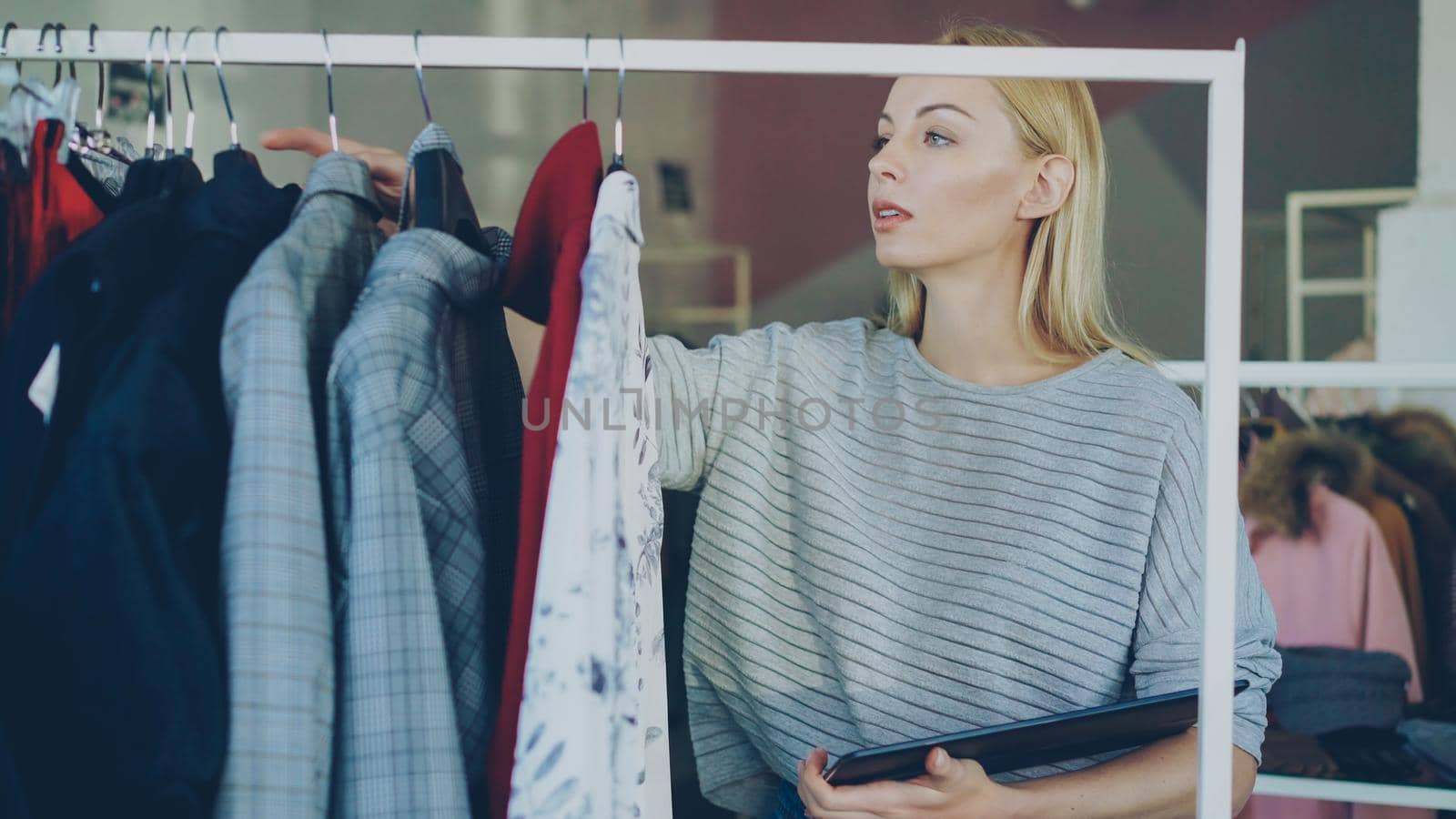 Owner of clothing store is checking and counting garments on rails while working with tablet. She is inputting information about her goods. Small business concept. by silverkblack