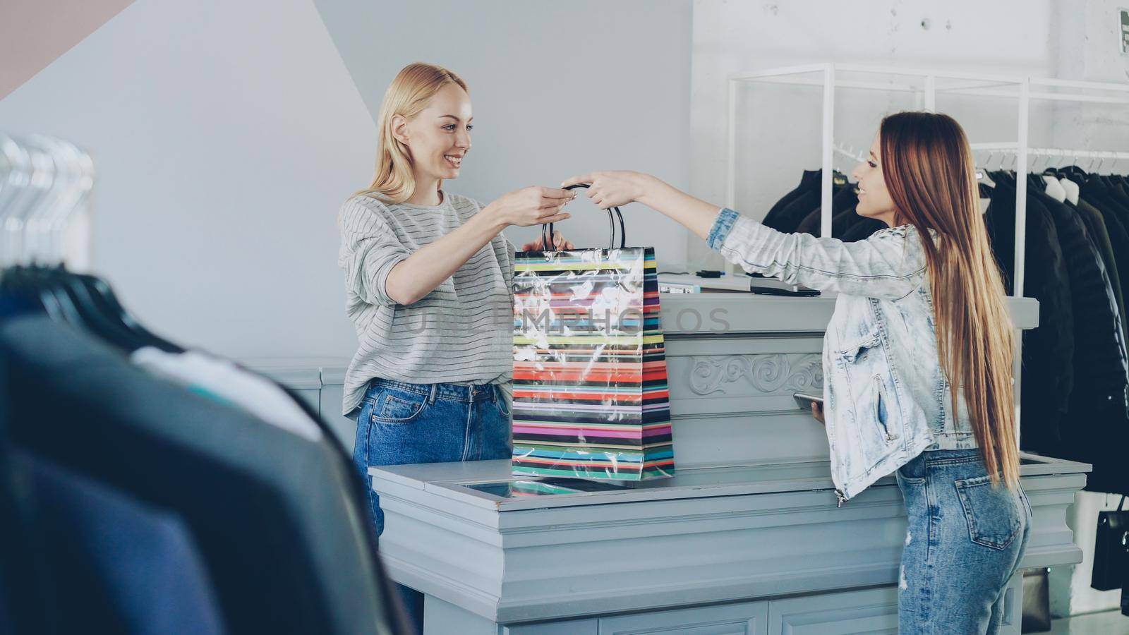 Cheerful customer purchased clothes while standing at cashier's desk. Shop assistant is accepting giving her paper bag by silverkblack