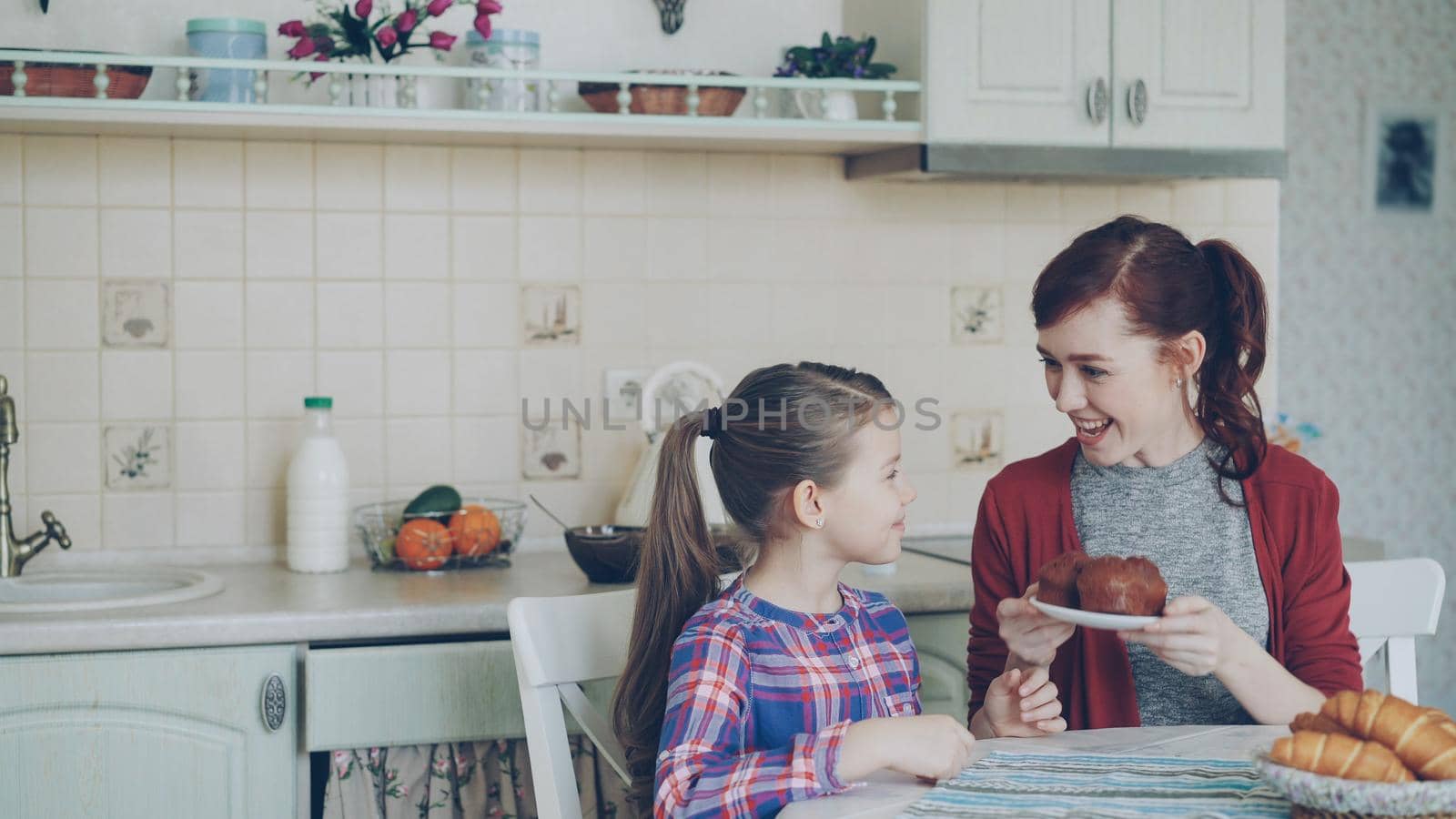 Young happy mother bringing tasty muffins to her cute daughter sitting at the table in kitchen waiting for breakfast at home