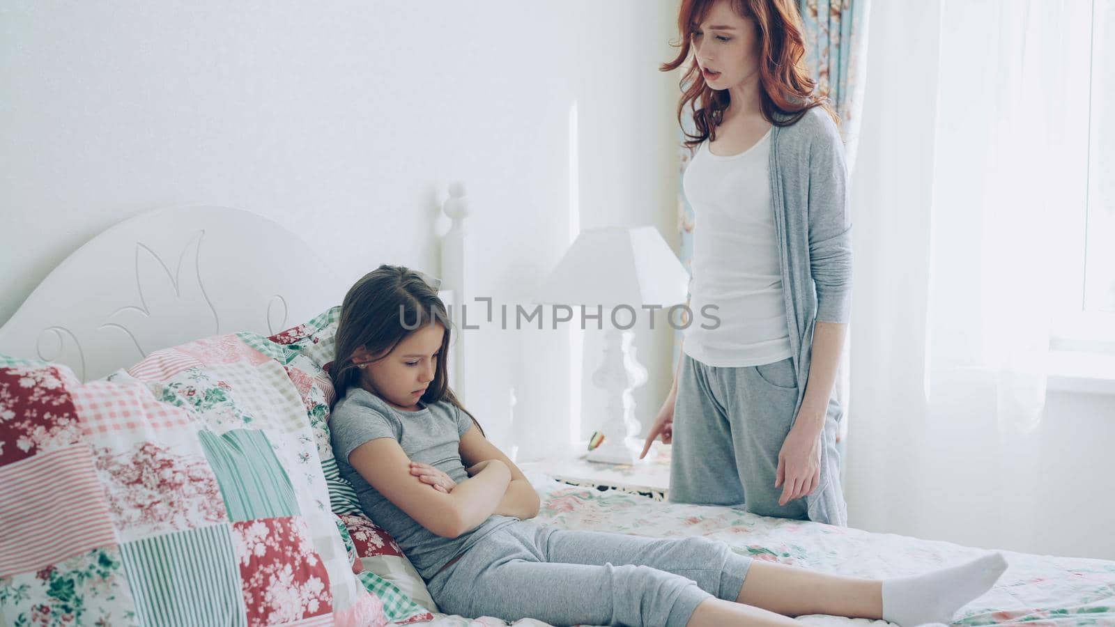 Angry mother argue scolding her upset little daughter lying on bed in bright bedroom at home by silverkblack