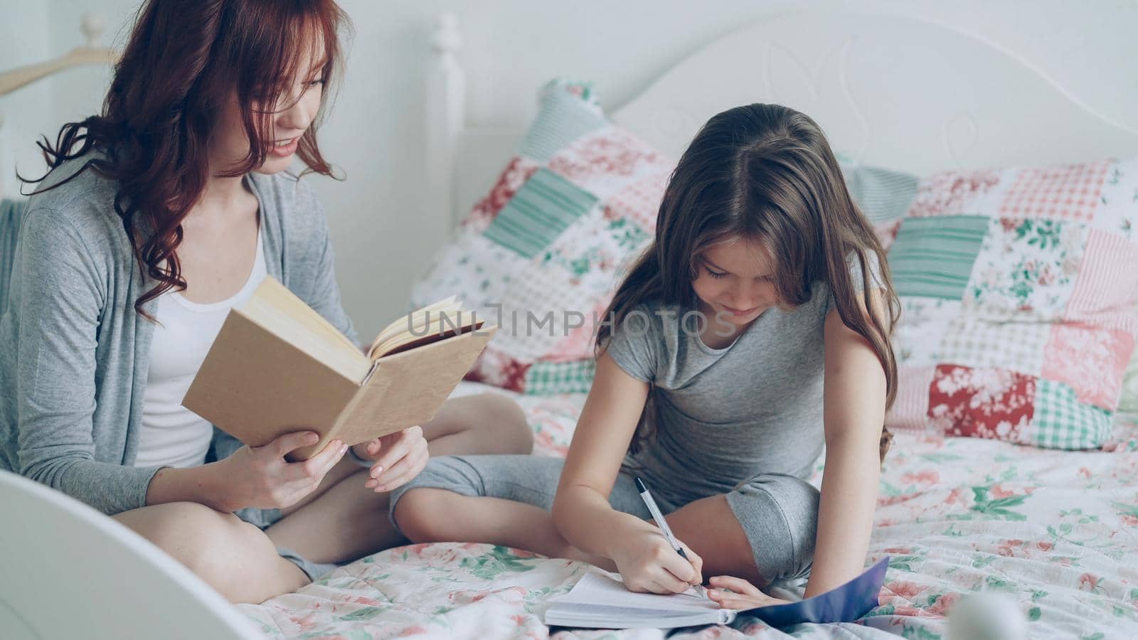 Careful mother helping her little cute daughter with homework for elementary school. Loving mom reading a book and girl writing notes in copybook while sitting together on bed at home by silverkblack