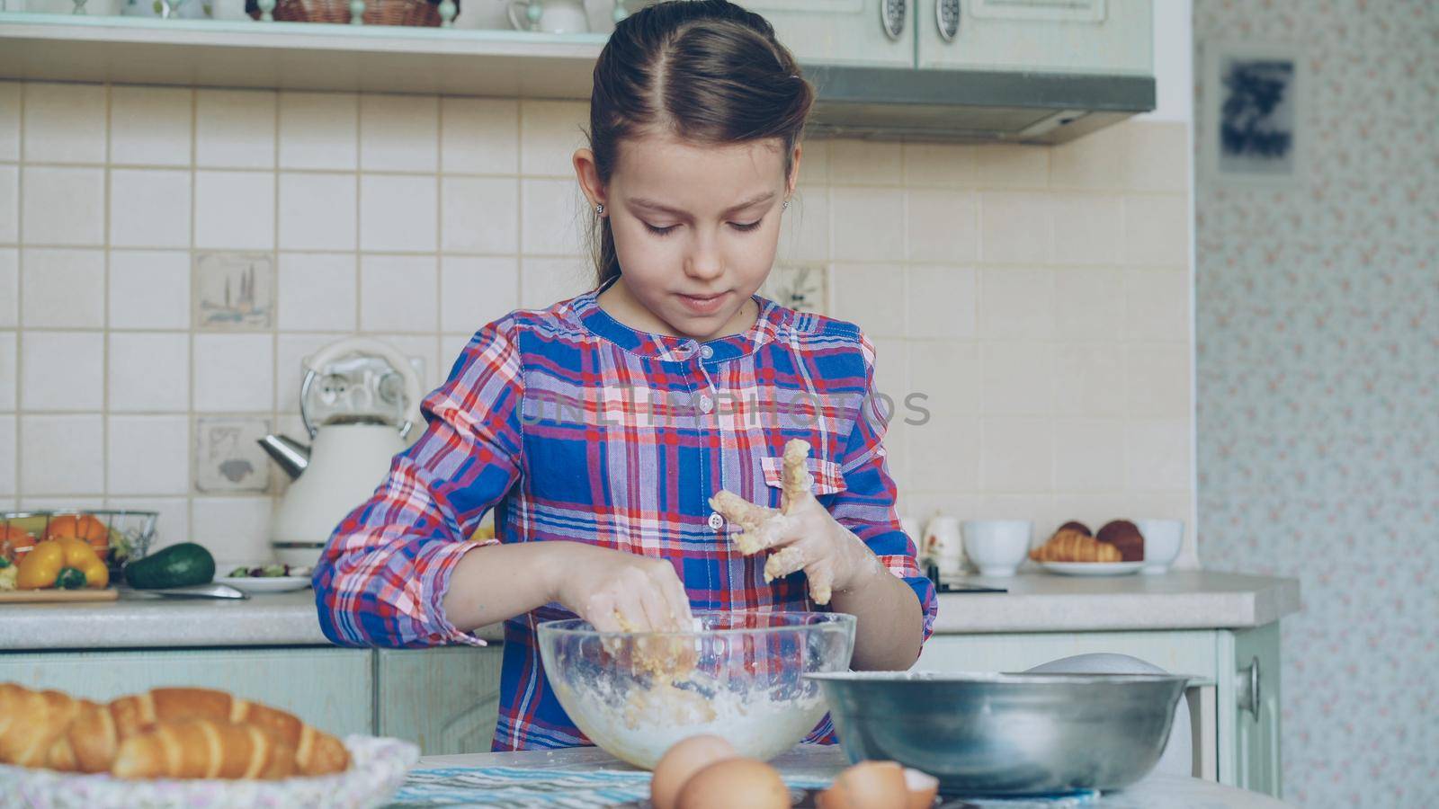 Little girl cooking dough and cleaning hands smiling in the kitchen at home. She wants to suprise her mother with baked cookies