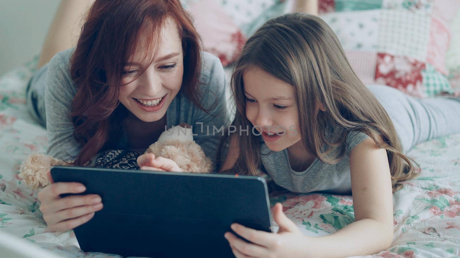 Close-up of smiling cute girl and young cheerful mother in pajamas laughing and looking in digital tablet while lying on bed at home