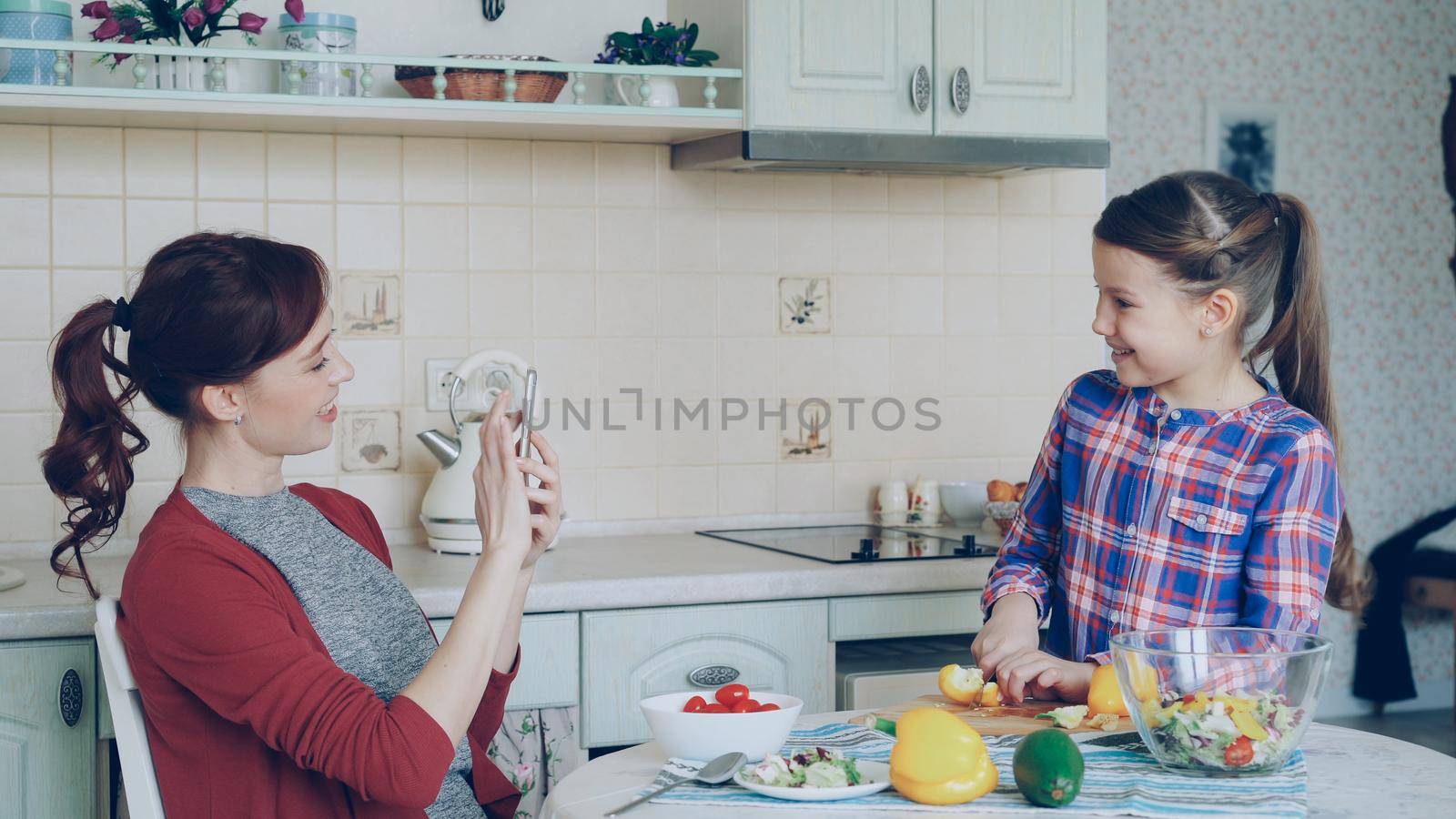 Young mother talking photo on smartphone camera of her cute smiling daughter cooking cutting vegetables in the kitchen at home. Family, cook, and people concept by silverkblack