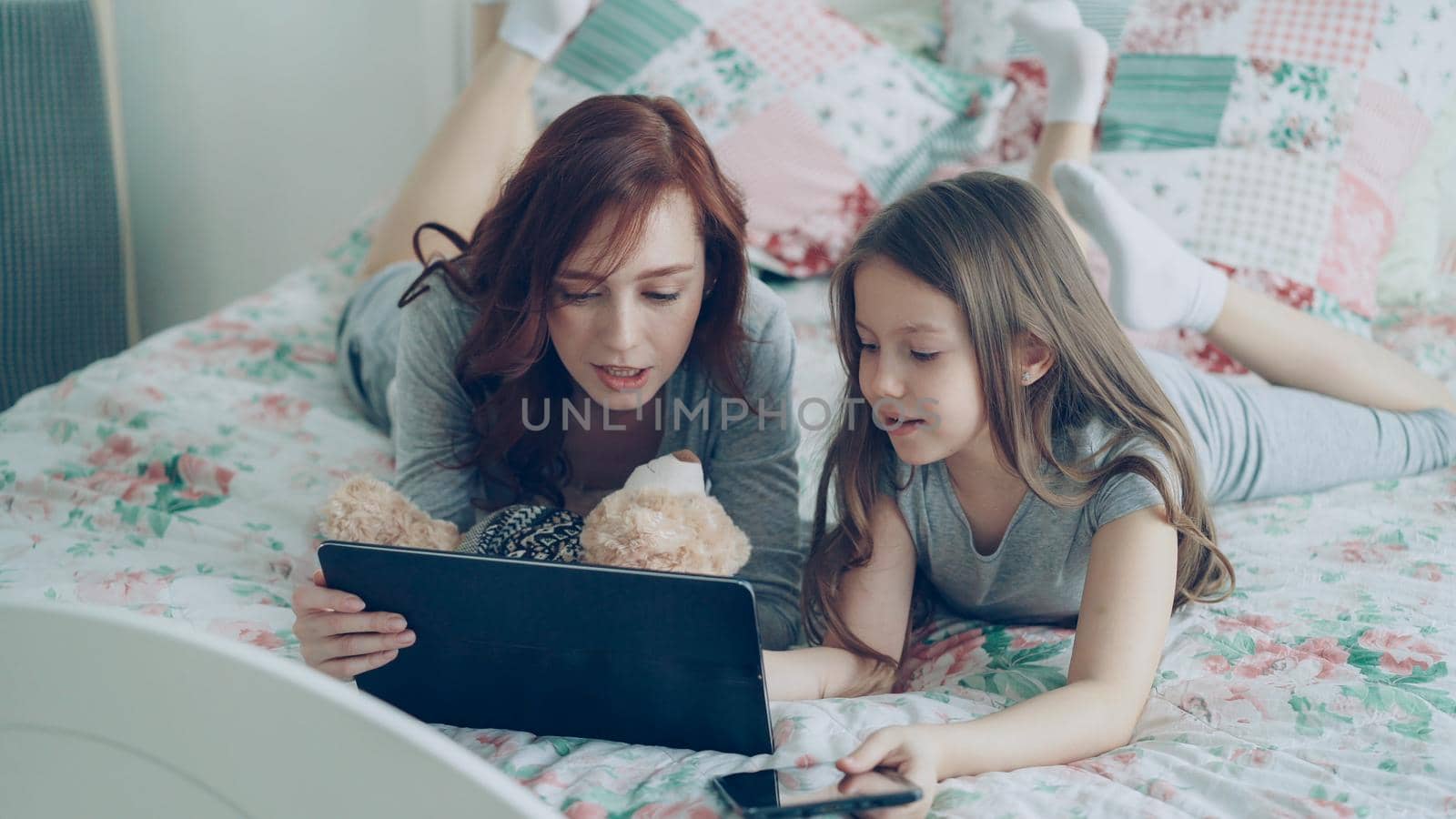 Beautiful young mother and her cute daughter in pajamas laughing and looking in digital tablet while lying on bed at home in the morning by silverkblack