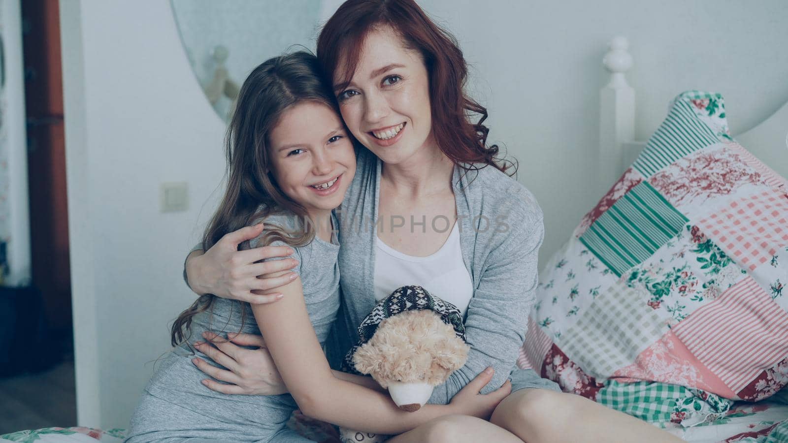Portrait of adorable smiling girl embracing her happy mother and looking at camera together while sitting on bed in bright cozy bedroom at home