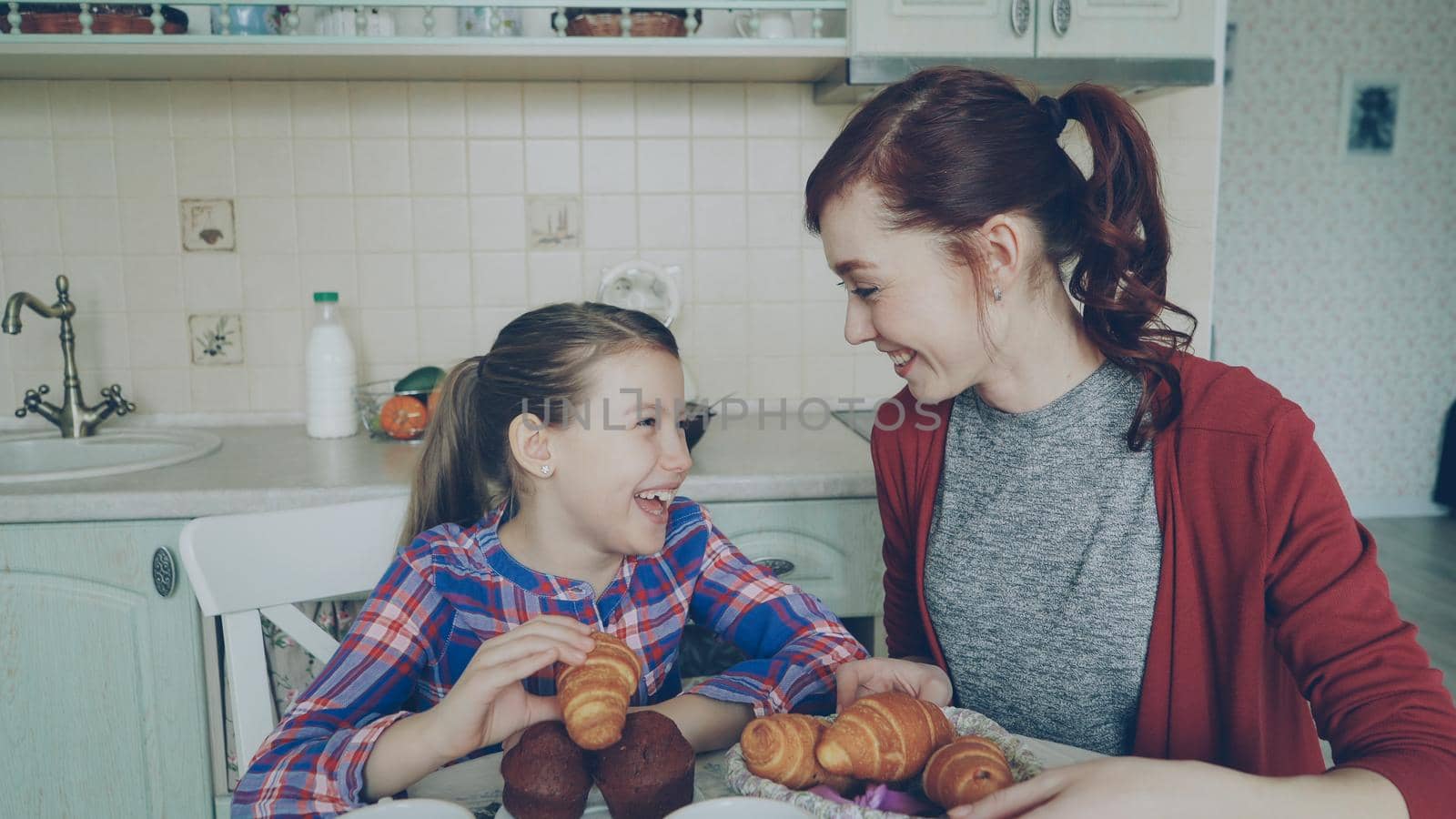 Cheerful mother and cute daughter having breakfast eating muffins and croissants talking at home in modern kitchen. Family, food, home and people concept by silverkblack