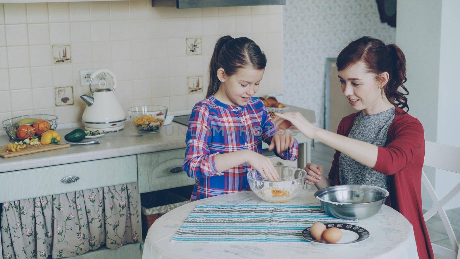 Little funny girl helping her mother in the kitchen cropping egg into bowl and mixing dough for cookies. Family, food, home and people concept by silverkblack