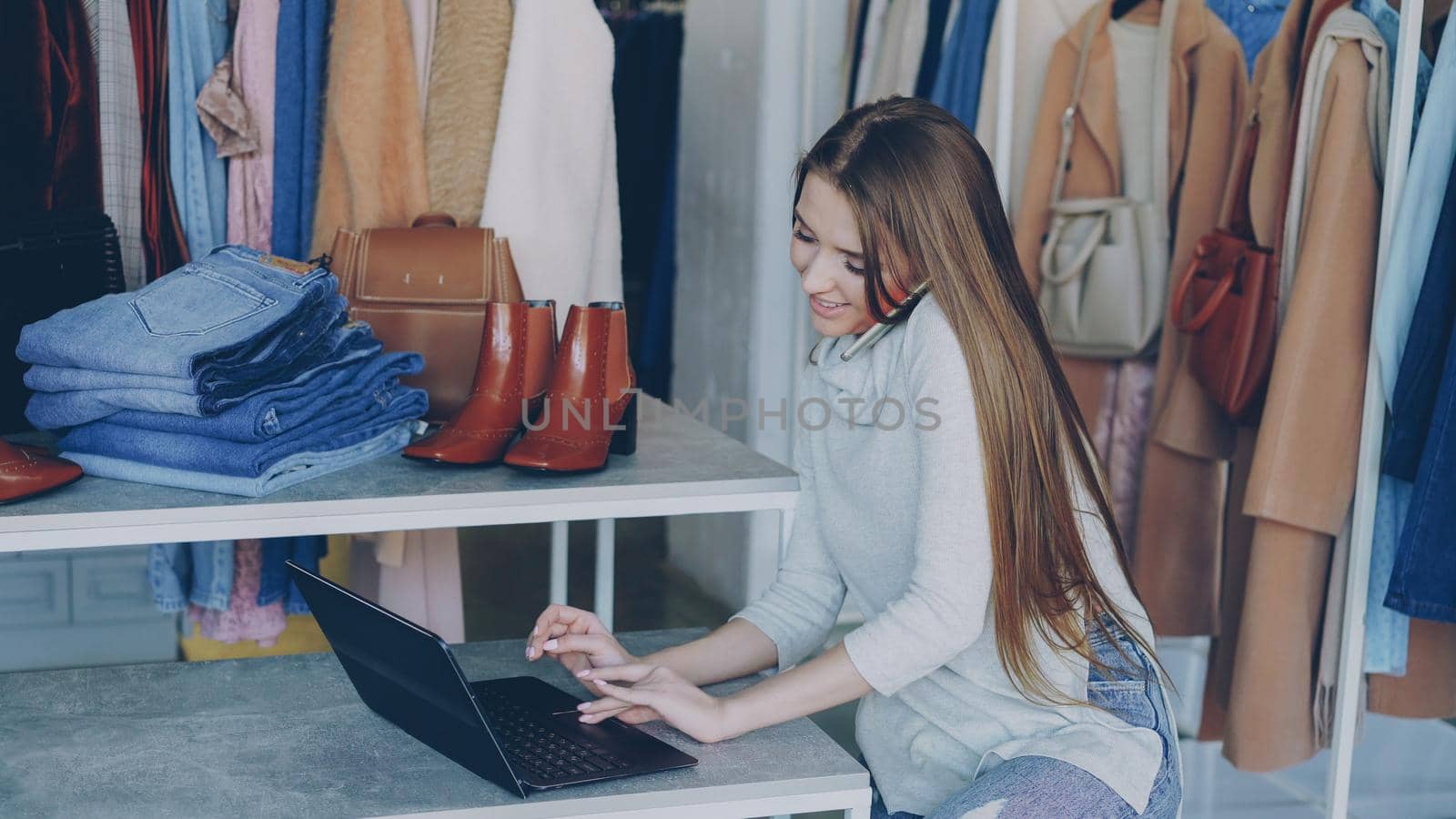 Attractive young businesswoman is sitting and talking on mobile phone while working with laptop in her women's clothing store. Colourful trendy garments, shoes and bags in background.