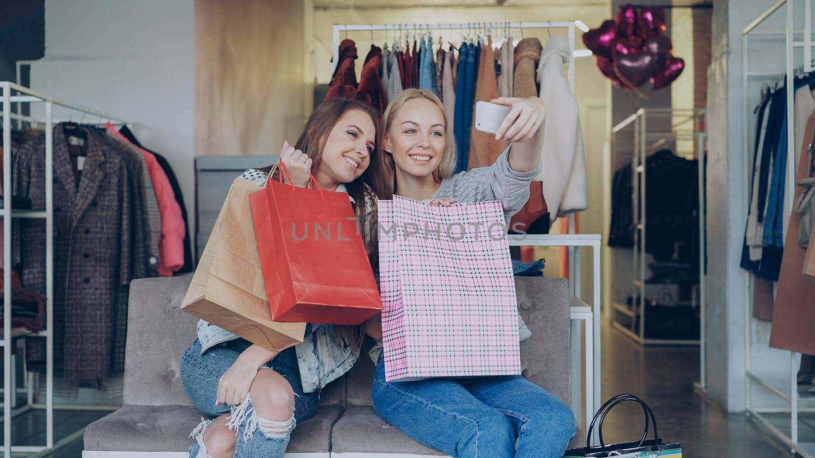 Attractive female friends are using smartphone to make selfie while sitting in women's clothing shop with lots of colourful paper bags. They are smiling, posing, laughing, gesturing.